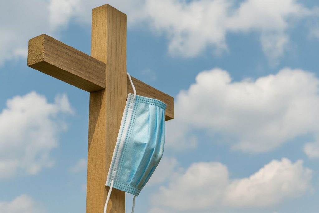 A wooden cross with a blue face mask symbolizes the psychological impact of COVID on faith and community.